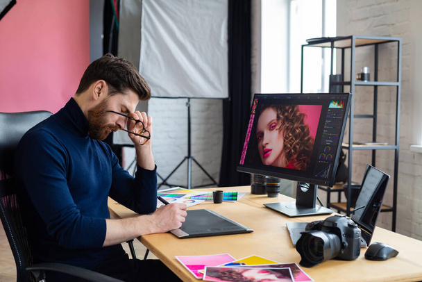 Portrait of tired graphic designer working overtime in office. Stressed worker have eye strain symptoms. Retoucher workplace in photo studio. Burnout and overwork concept. Creative agency - 写真・画像
