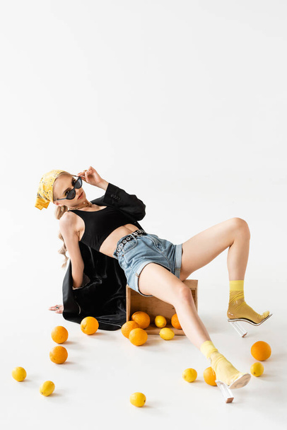 fashionable woman posing on wooden box near scattered citrus fruits on white background - Photo, Image