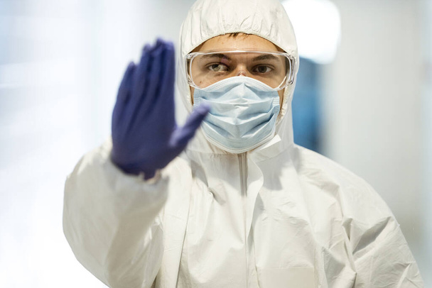 Coronavirus-Konzept. Männlicher Wissenschaftler Arzt mit Maske, Brille und Schutzanzug, der während des Ausbruchs der COVID-Pandemie ein Stoppschild mit seiner Hand in Latexhandschuhen zeigt. Der Mensch im biochemischen Labor. - Foto, Bild