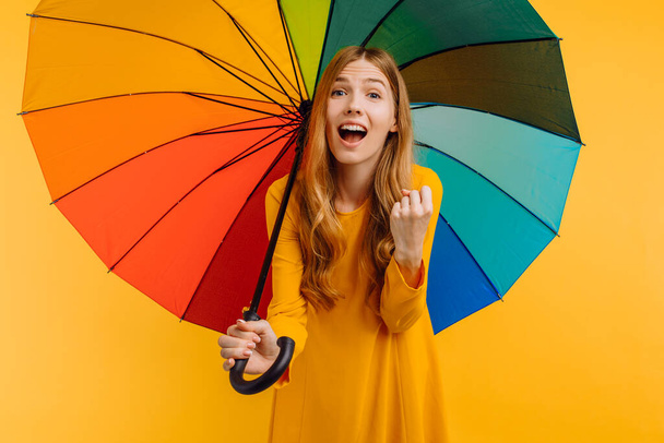 Shocked surprised girl, in a yellow dress, happy and posing with a rainbow umbrella, showing a winning gesture on an isolated yellow background - Zdjęcie, obraz
