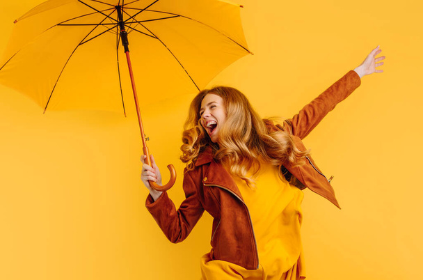 funny girl in a yellow dress and autumn jacket, laughing and jumping with a yellow umbrella on a yellow background - Foto, afbeelding