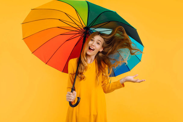 Sarı elbiseli mutlu, çekici bir kız, izole edilmiş sarı bir arka planda gökkuşağı şemsiyesiyle eğleniyor. - Fotoğraf, Görsel