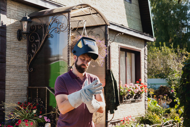 βάναυση γενειοφόρος συγκολλητής φορώντας μάσκα προσώπου και προστατευτικά γάντια που εργάζονται με μια μηχανή συγκόλλησης σε ένα οικόπεδο εξοχή μπροστά από το σπίτι καλοκαιρινή μέρα  - Φωτογραφία, εικόνα