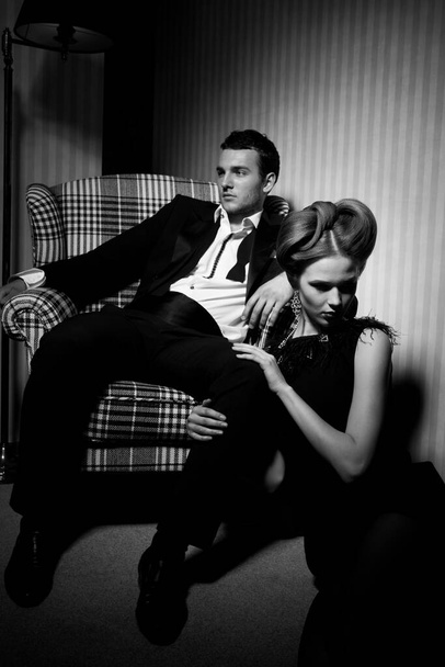 Hermosa pareja apasionada en ropa clásica en las sombras de la habitación: hombre en la silla, mujer en el suelo a sus pies - Foto, imagen