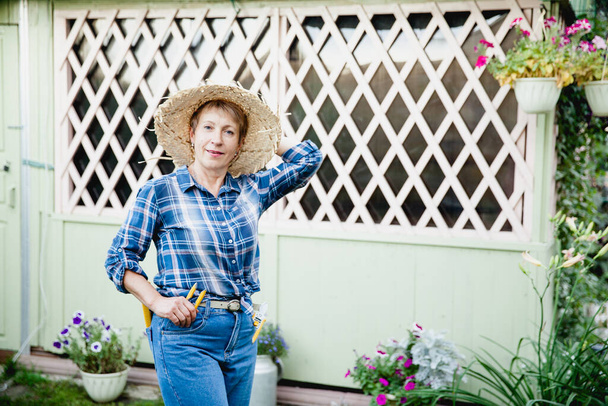 стильная пожилая женщина садовница в синих джинсах, клетчатая рубашка и соломенная шляпа на фоне деревянной беседки - Фото, изображение