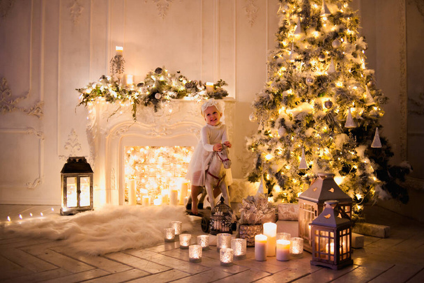 adorable kid in white dress on ride on horse toy near Christmas tree with garlands and fireplace - Photo, Image
