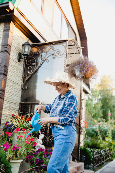 όμορφη ευτυχισμένη γυναίκα συνταξιούχος κηπουρός σε τζιν, ένα πουκάμισο σε ένα κλουβί και ένα ψάθινο καπέλο στέκεται σε ένα φόντο ενός εξοχικού σπιτιού με ένα μπλε ποτιστήρι στα χέρια της πότισμα λουλούδια - Φωτογραφία, εικόνα