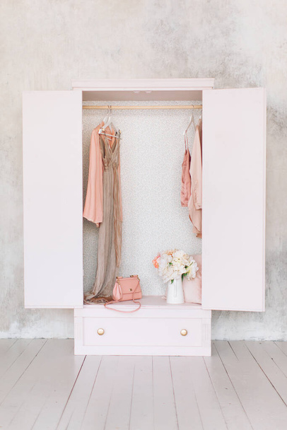 wooden pink wardrobe for women clothes, open doors, decor, mirror, bag, hangers, silk dresses in a bright room, concept, flowers delicate pastel colors - Photo, Image
