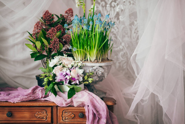 vintage décoré avec des fleurs de printemps en tissu violet et des verts sur des meubles anciens en bois avec tiroirs dans une chambre classique sur un plancher en bois blanc près de grandes fenêtres et rideaux en tulle - Photo, image
