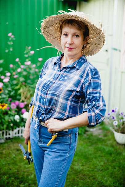 stylowa kobieta emerytowany ogrodnik w niebieskich dżinsach, koszuli w kratkę i słomy kapelusz pobyt na tle drewnianej altanki - Zdjęcie, obraz