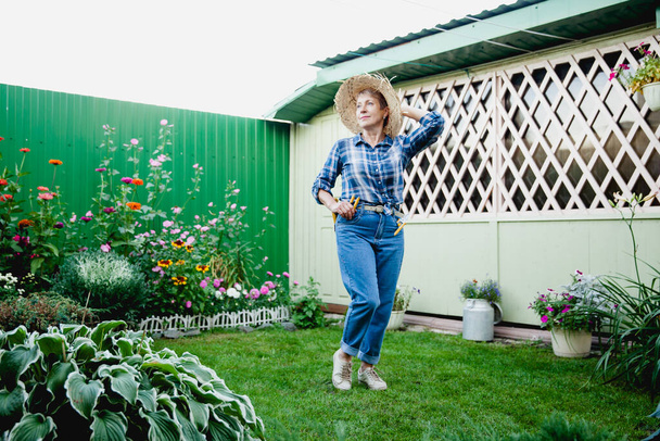Stilvolle alte Gärtnerin im Ruhestand in blauen Jeans, kariertem Hemd und Strohhut auf dem Hintergrund einer hölzernen Gartenlaube - Foto, Bild