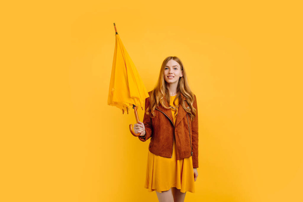 joyous beautiful girl, in a bright yellow dress and autumn jacket, stands with a yellow umbrella on an isolated yellow background - Zdjęcie, obraz