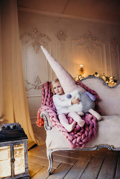 αξιολάτρευτο παιδί σε λευκό φόρεμα στον καναπέ με κουβέρτα βελονάκι και μεγάλο παιχνίδι, Χριστουγεννιάτικες γιρλάντες στο στούντιο - Φωτογραφία, εικόνα