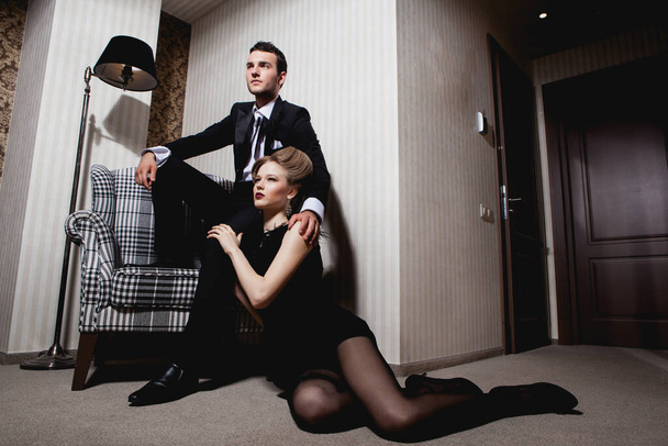 Belo casal apaixonado em roupas clássicas nas sombras da sala: homem na cadeira, mulher no chão a seus pés - Foto, Imagem