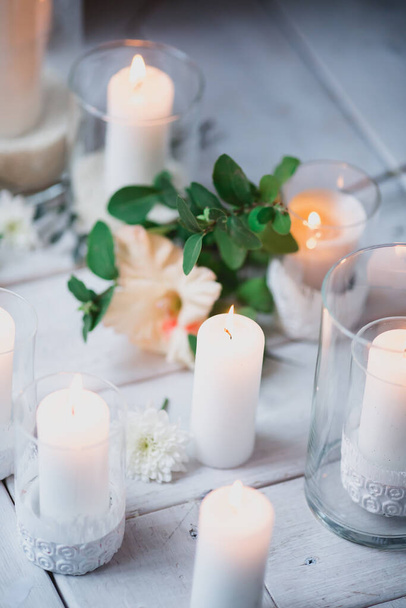 piękny wystrój białych świec w szklanych świeczników stoją na białej drewnianej podłodze wśród zielonych liści i kwiatów - Zdjęcie, obraz