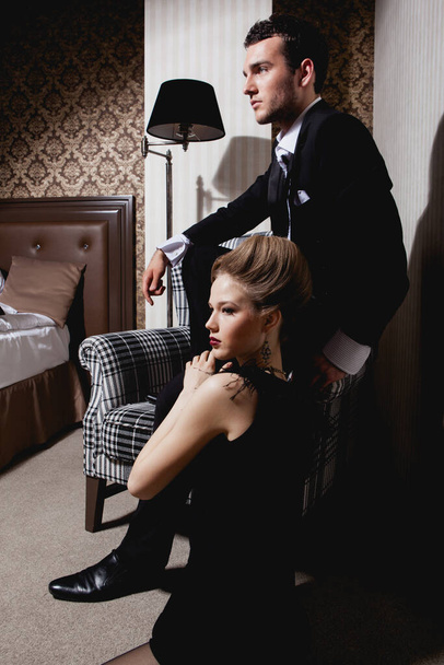Gyönyörű szenvedélyes pár klasszikus ruhában a szoba árnyékában: férfi a széken, nő a földön a lábainál - Fotó, kép