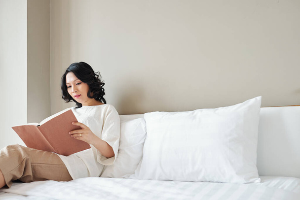 Pretty Asian woman sitting on bed and reading interesting book - Photo, Image