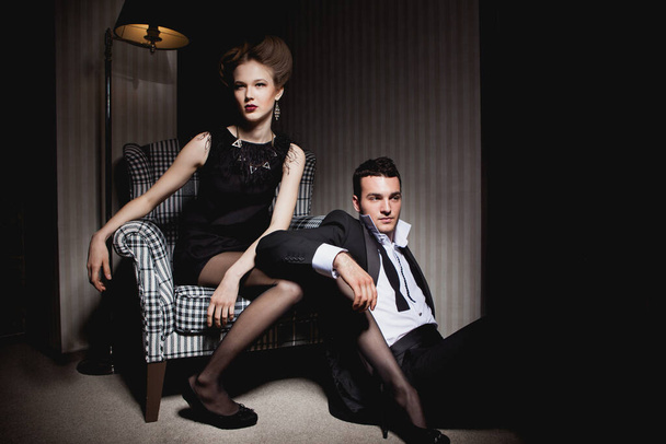 Hermosa pareja apasionada en ropa clásica en las sombras de la habitación: mujer en silla, un hombre en el suelo a sus pies - Foto, imagen