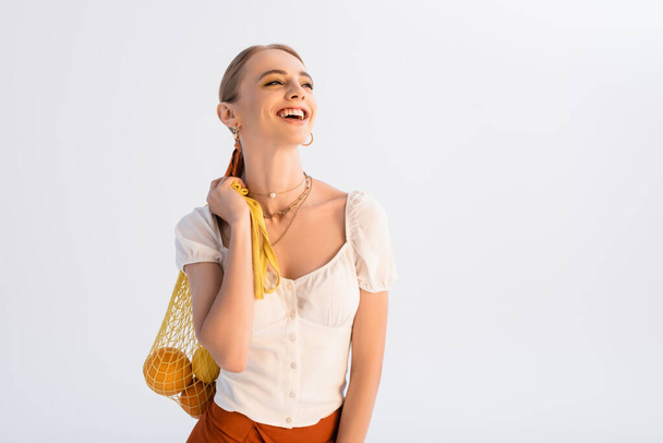 rustic blonde woman with citrus fruits in yellow string bag laughing isolated on white - 写真・画像