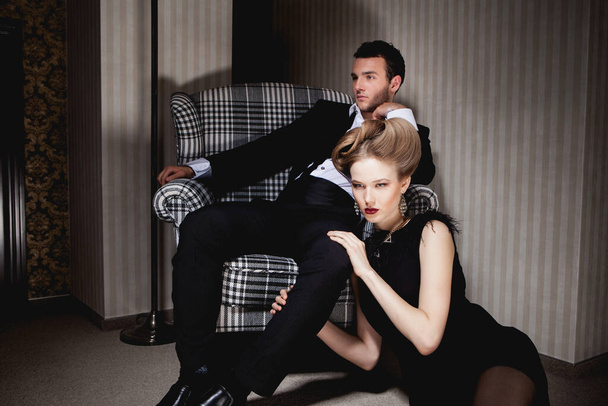 Mooi gepassioneerd koppel in klassieke kleding in de schaduwen in de kamer: man op de stoel, vrouw op de vloer aan zijn voeten - Foto, afbeelding