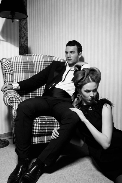 Hermosa pareja apasionada en ropa clásica en las sombras de la habitación: hombre en la silla, mujer en el suelo a sus pies - Foto, imagen