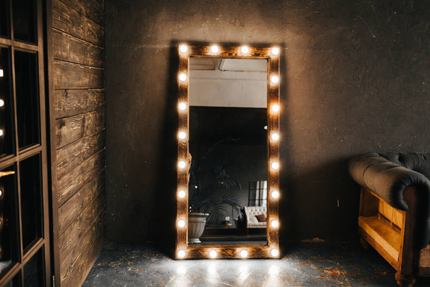 Corner room with a wooden and gray cement wall, an aged floor, black glass doors and electric dressing makeup mirror with lights bulbs and oxford sofa in loft style - Φωτογραφία, εικόνα