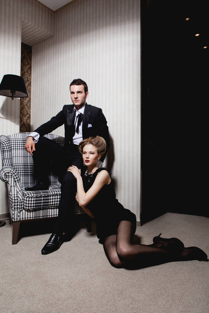 Mooi gepassioneerd koppel in klassieke kleding in de schaduwen in de kamer: man op de stoel, vrouw op de vloer aan zijn voeten - Foto, afbeelding
