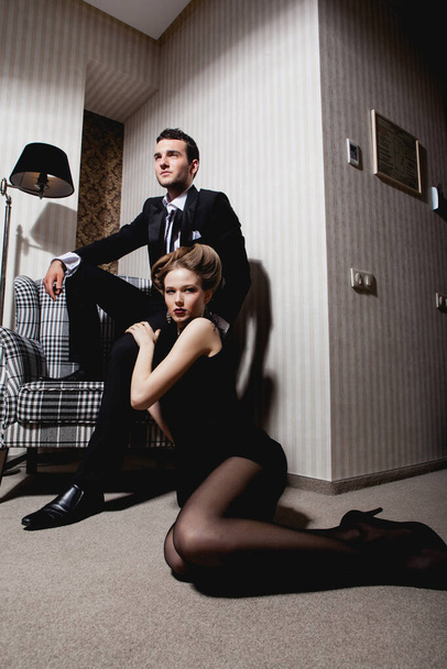 Bella coppia appassionata in abiti classici nell'ombra nella stanza: uomo sulla sedia, donna sul pavimento ai suoi piedi - Foto, immagini