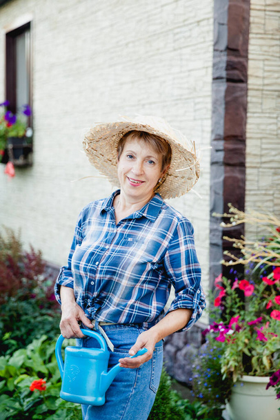mooie vrolijke vrouw oud gepensioneerde tuinman in spijkerbroek, een shirt in een kooi en een strohoed staat op de achtergrond van een landhuis met een blauwe gieter in haar handen water geven bloemen - Foto, afbeelding