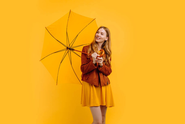 cheerful girl in a yellow dress and autumn jacket, laughing and rejoicing with a yellow umbrella on a yellow background - Photo, Image