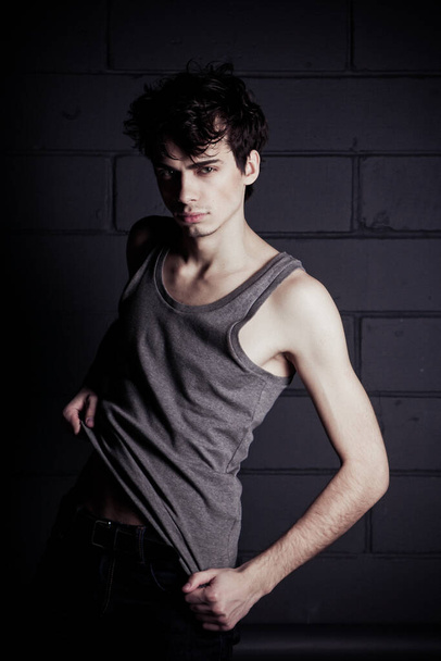 A young thin dark-haired guy in a gray T-shirt posing against a concrete-brick wall - Foto, immagini