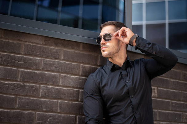 Portrait of a charming young businessman dressed in suit standing outside a glass building and touching his sunglasses - Foto, imagen