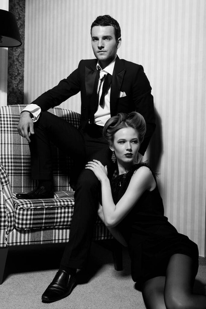 Hermosa pareja apasionada en ropa clásica en las sombras de la habitación: hombre en la silla, mujer en el suelo a sus pies - Foto, Imagen