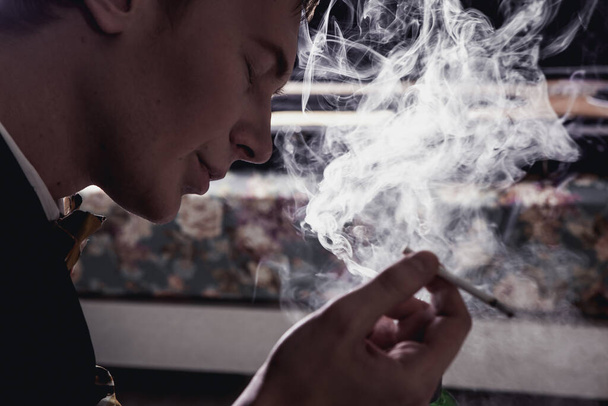 handsome man in black jacket sitting and smoking in room - Zdjęcie, obraz