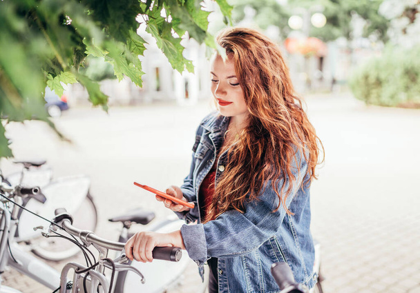 Portrait of smiling red curled long hair caucasian teen girl unlocking bike at Bicycle sharing point using the modern smartphone. Green urban transport and modern technology devices concept image. - Фото, зображення