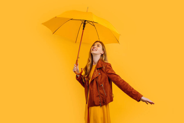 Happy cheerful girl in a yellow dress and autumn jacket, laughing and having fun with a yellow umbrella on a yellow background, concept of autumn - Photo, image