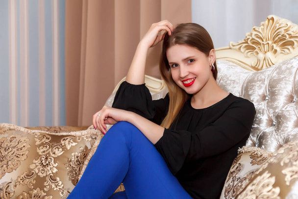 Woman on sofa of hotel room. Pretty woman of Slavic appearance in black blouse and blue pants. Happy lady. Spend time on vacation. Cute girl is smiling and shows emotions - Photo, image