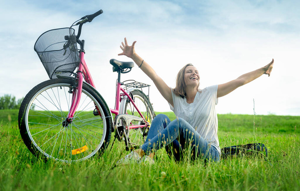 Happy woman sitting on the grass in a green meadow next to the bike and raising her hands up, summer vacation and adventure concept. - Fotoğraf, Görsel