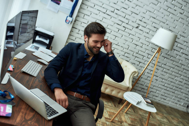 Young successful businessman or sales manager wearing wireless earphones, talking with client or partner and smiling while working in the modern office - Valokuva, kuva