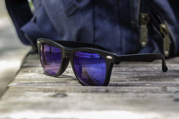 Sunglasses lie on a wooden park bench. Blue backpack in the background. Hiking or traveling. Active lifestyle. Without people. Selective focus. - Photo, Image