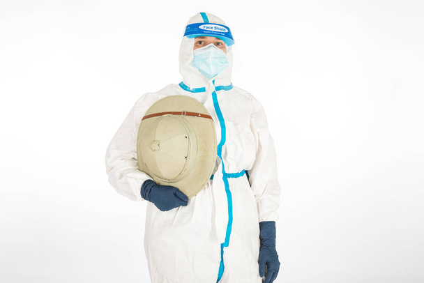 A doktornő PPE-t visel, hogy harcoljon a koronavírus ellen, kezében egy gyarmati korszak felfedező kalapját tartva, fehér háttérrel. Harcos. COVID gyógyszer koncepciója - Fotó, kép