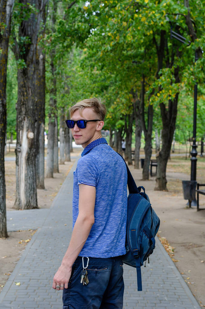 Portrait de jeune homme en lunettes de soleil et sac à dos derrière son dos dans le parc de la ville. Jeune homme en chemise bleue tourné en arrière et regardé la caméra. Mode de vie. - Photo, image