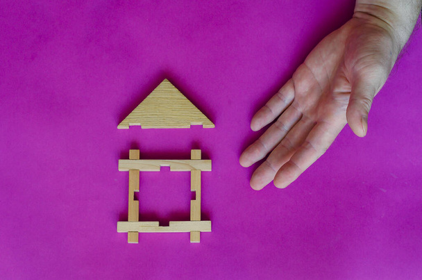 La main masculine pointe vers la maison en bois jouet sur fond lilas. Le concept de construction, d'hypothèque, de vente immobilière ou de prêt garanti par l'immobilier. Vue d'en haut. - Photo, image