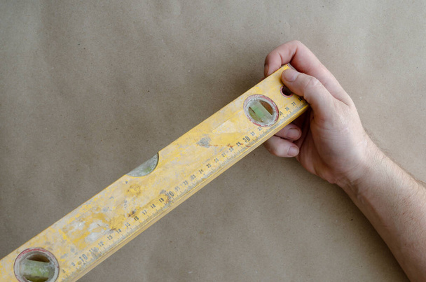 Hand of an adult Caucasian man holds a yellow construction level. A real hand tool with scratches and traces of cement. The construction industry. Selective trick. - Foto, afbeelding