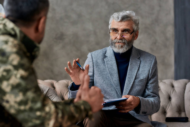 Fixing you. Portrait of mature psychologist communicating with military man and making notes during therapy session. Soldier suffering from depression, psychological trauma. PTSD concept - Photo, Image