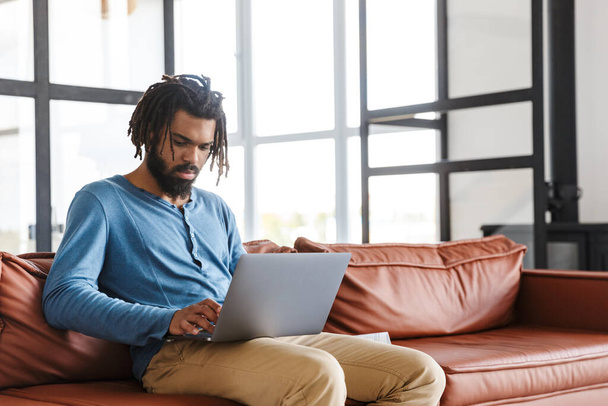 Handsome young african man sitting on a leather couch at home, using laptop computer - Foto, Imagen