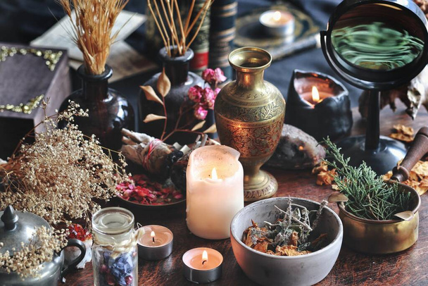Dried plants, herbs and yellow rose flower petals in a grey, dirty clay pot. Wiccan witch altar with ingredients on it ready to make cast a spell. Evergreens, books, burning candles in dark background - Photo, Image
