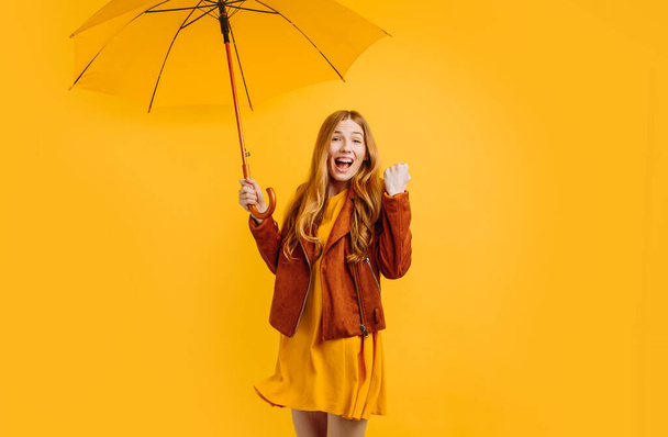 happy girl in a yellow dress and autumn jacket, with a yellow umbrella, is happy and shows a winning gesture on an isolated yellow background - Foto, afbeelding