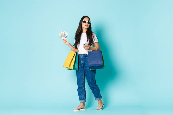 Mode, Schönheit und Lifestylekonzept. Freche junge Asiatin mit Sonnenbrille schaut sich beim Einkaufen um, hält Geld, Taschen mit Kleidung und Handy, hellblauer Hintergrund - Foto, Bild