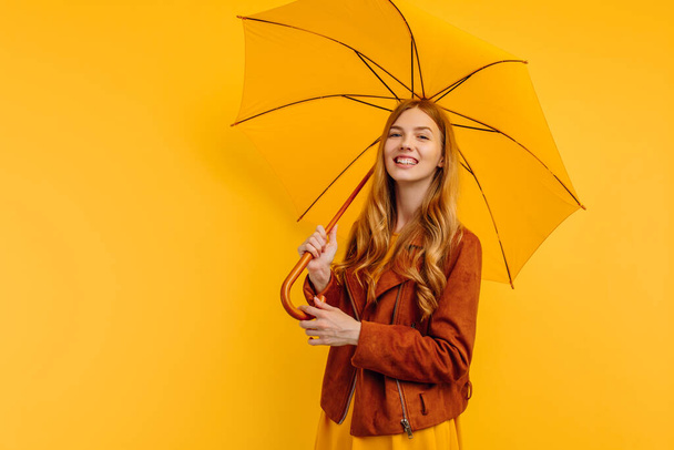 happy attractive girl, in a bright yellow dress and autumn jacket, stands with a yellow umbrella on an isolated yellow background - Photo, image
